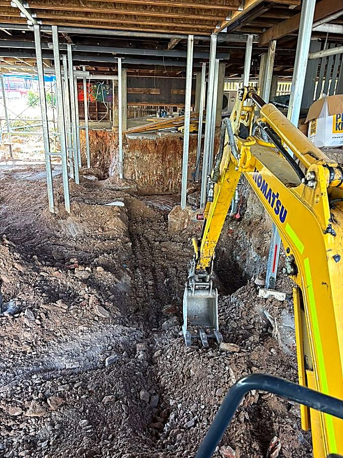 Excavator digging a plumbing trench under a house